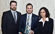 Photo of two men and one woman posing for a group picture. The man in the center is holding an award.