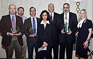 Photo of five men and two women posing for a group picture. Three of the men are holding awards.