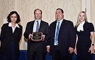 Photo of two women and two men posing for a group picture. One man in the center is holding an award.