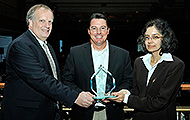 Photo of two men and one woman posing for a group picture. The man in the center is holding an award.