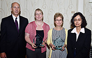 Photo of three women and one man posing for a group picture. The two women in the center are holding awards.