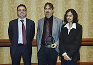 Photo of two men and one woman posing for a picture. The man in the center is holding an award.