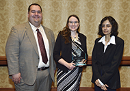 Photo of one man and two women posing for a picture. The woman in the center is holding an award.