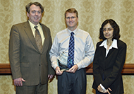 Photo of two men and one woman posing for a picture. The man in the center is holding an award.