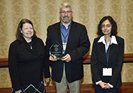 Photo of two women and one man posing for a picture. The man in the center is holding an award.