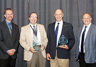 Photo of four men posing for a group picture. Two men in the center are holding awards.