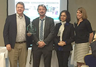 Photo of two men and two women posing for a group picture. One man on the left is holding an award.