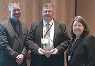 Photo of two men and one woman posing for a group picture. The man in the center is holding an award.