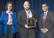 Photo of one woman and two men posing for a group picture. The man in the center is holding an award.