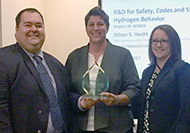 Photo of one man and two women posing for a group picture. The woman in the center is holding an award.