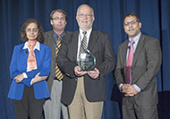 Photo of three men and one woman posing for a group picture. One man in the center is holding an award.