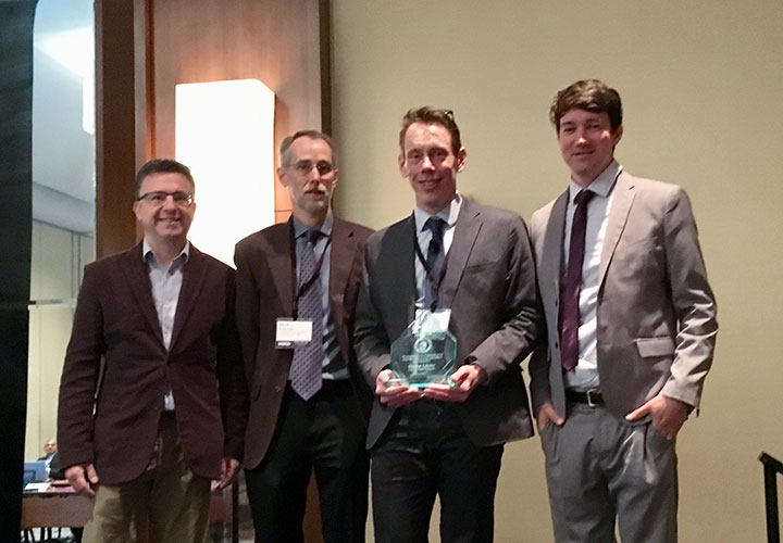 Four men, standing and smiling in a conference room, one holding an award