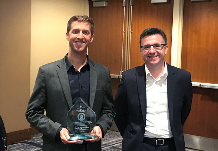 Two men, standing and smiling in a conference room, one holding an award