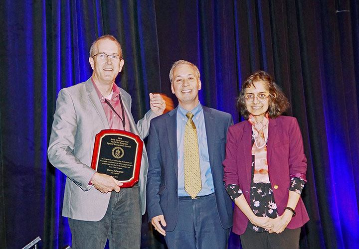 Two men and one woman, standing and smiling in a conference room, one of the men holding an award