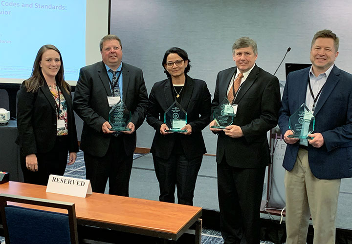 Two women and three men, standing and smiling in a conference room, all but one of them holding awards