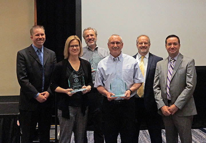 One woman and four men, standing and smiling in a conference room, one of the men and one of the women holding awards