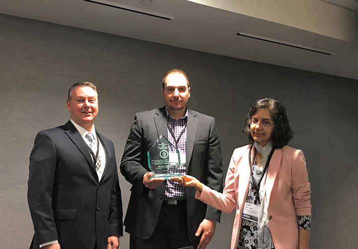 Two men and one woman standing and smiling in a conference room, one of the men holding an award