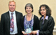 Photo of one man and two women posing for a group picture. One of the women is holding an award.