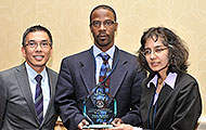 Photo of two men and one woman posing for a group picture. One of the men is holding an award.