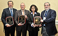 Photo of three men and one woman posing for a group picture. The three men are holding award plaques.