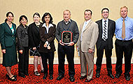 Photo of four women and four men posing for a group picture. The man in the center is holding an award plaque.